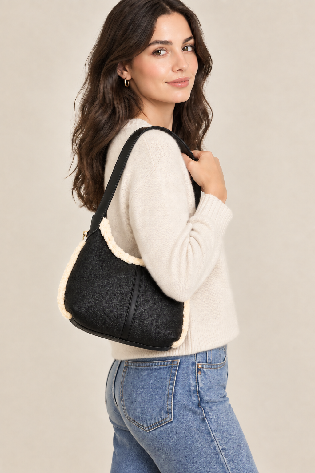 A woman wearing a cream knit sweater and blue jeans carries a black shoulder bag with cream sherpa lining and trim, photographed against a plain beige studio background.