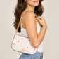 A woman wearing a white tank top and blue jeans carries a small white Kate Spade crossbody bag with a gold chain strap and red and pink heart embellishments against a light beige studio background.