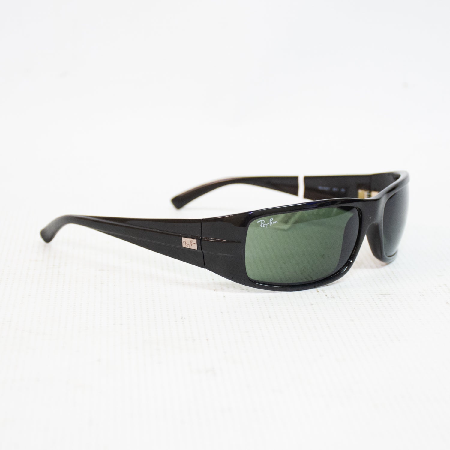 Ray-Ban wraparound sunglasses with a glossy black acetate frame and dark green lenses, displayed on a white textured surface.